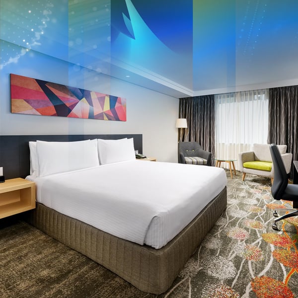 A hotel room with a king bed, colourful artwork above it and armchairs by the window